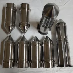 Srebrne, metalowe elementy o stożkowych zakończeniach, wykonane z precyzją, ułożone na białym tle, niektóre w pozycji pionowej, inne poziome, ukazujące detale obróbki.