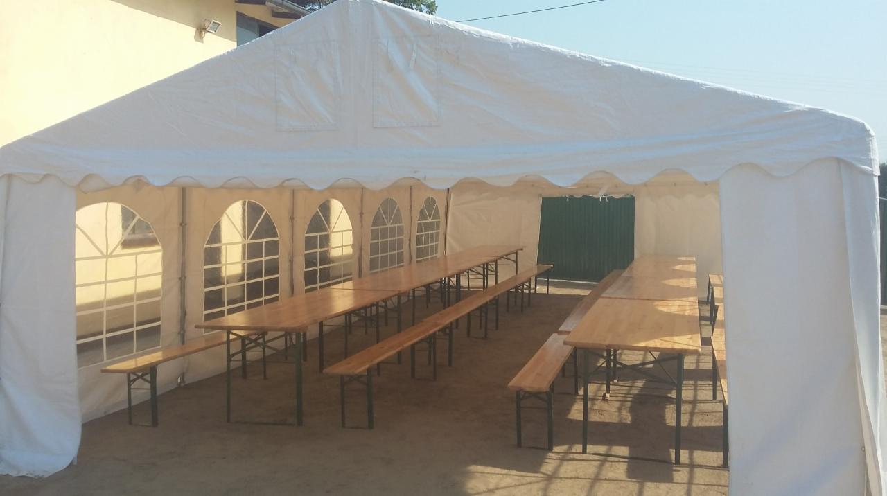 Wnętrze białego namiotu imprezowego z drewnianymi stołami i ławkami, widok z zewnątrz w słoneczny dzień.