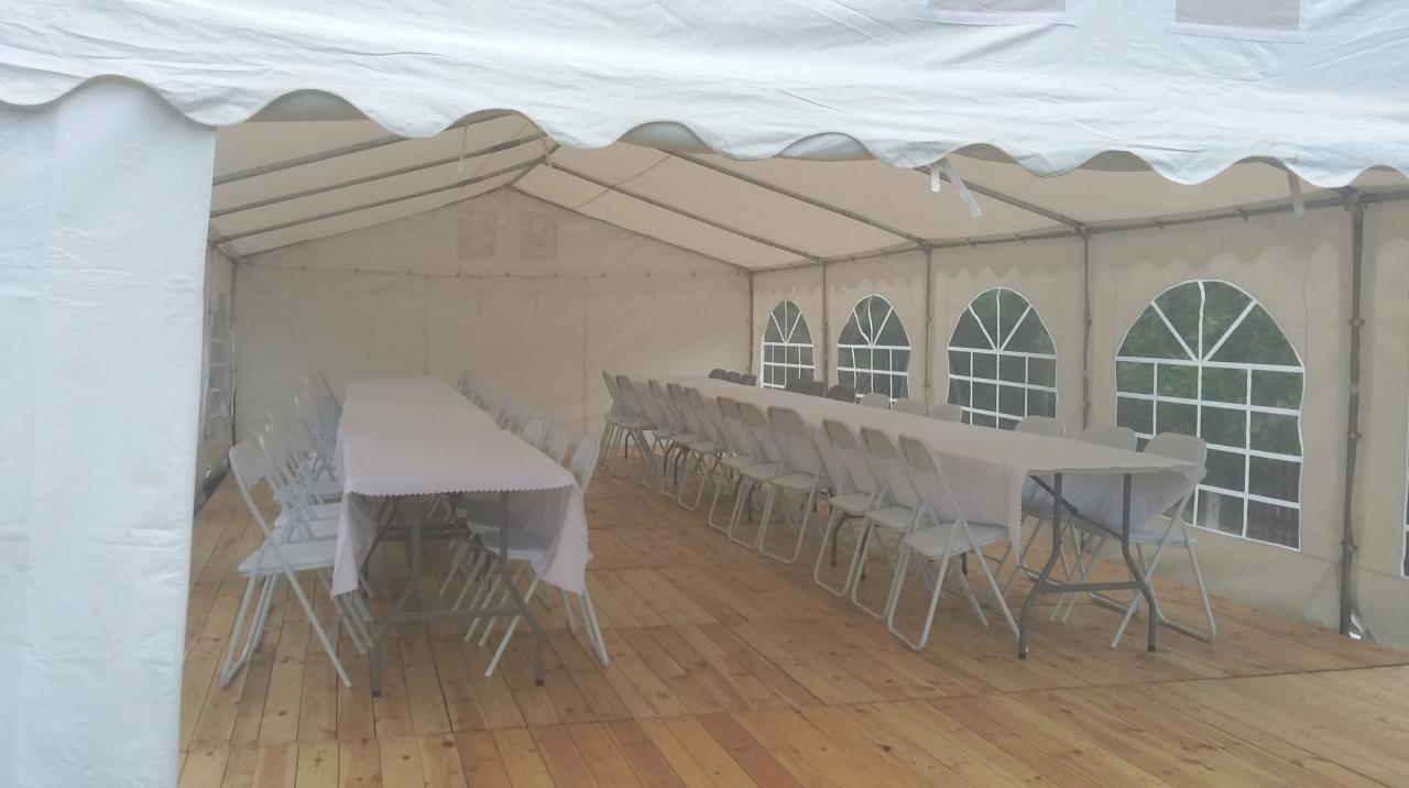 Wnętrze białego namiotu imprezowego z drewnianą podłogą, ustawione stoły z białymi obrusami i krzesłami, widoczne okna z widokiem na zieleń.