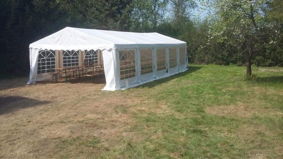 Rozłożony biały namiot imprezowy z łukowatymi oknami, ustawiony na trawniku z widocznymi stołami i ławkami wewnątrz.