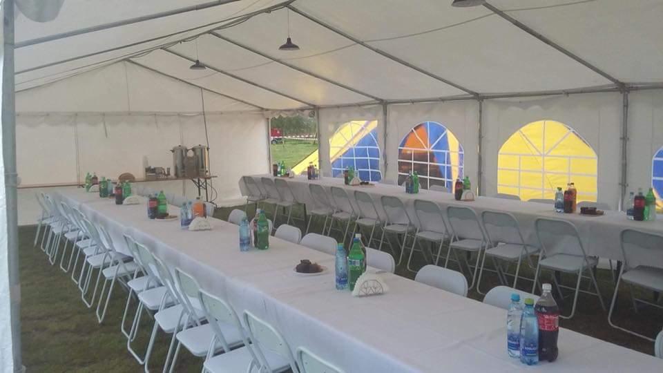 Wnętrze białego namiotu imprezowego z długim stołem nakrytym białym obrusem, ustawionymi krzesłami, napojami i dekoracjami; widoczne kolorowe okna namiotu oraz sprzęt cateringowy w tle.