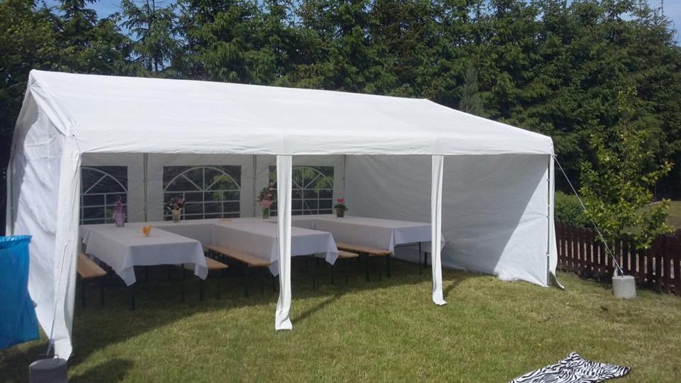 Biały namiot imprezowy rozstawiony na trawie, z trzema stołami nakrytymi białymi obrusami i drewnianymi ławkami wewnątrz, dekoracje kwiatowe na stołach, widok z boku, w tle zielone drzewa i brązowe...