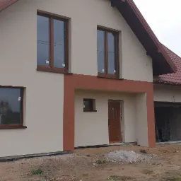 Nowo wybudowany dom jednorodzinny z beżową elewacją, brązowymi oknami i dachem, widok na garaż i niezagospodarowany teren wokół