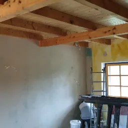 Ściana po nałożeniu gładzi szpachlowej, widoczne drewniane belki stropowe, fragment drabiny i osoba w tle, pomieszczenie w trakcie remontu.