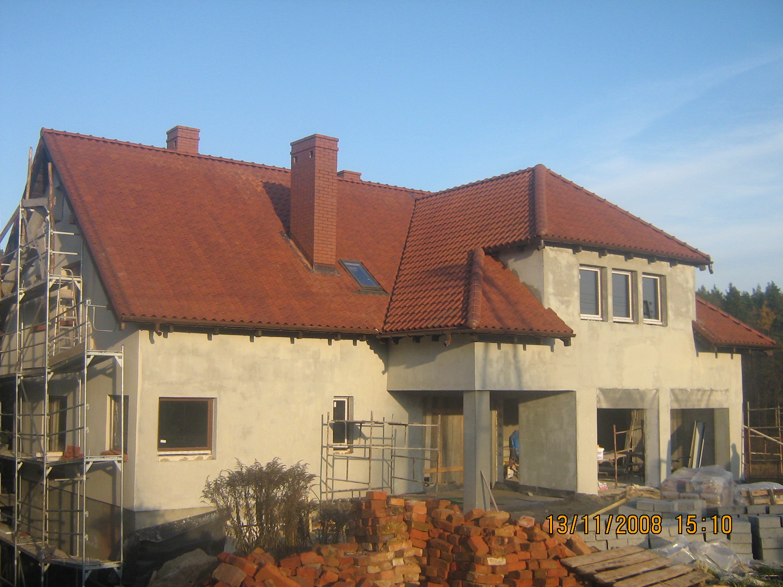 Dom w trakcie budowy z czerwoną dachówką, widoczne rusztowania, stos cegieł i bloczków oraz data na pierwszym planie.