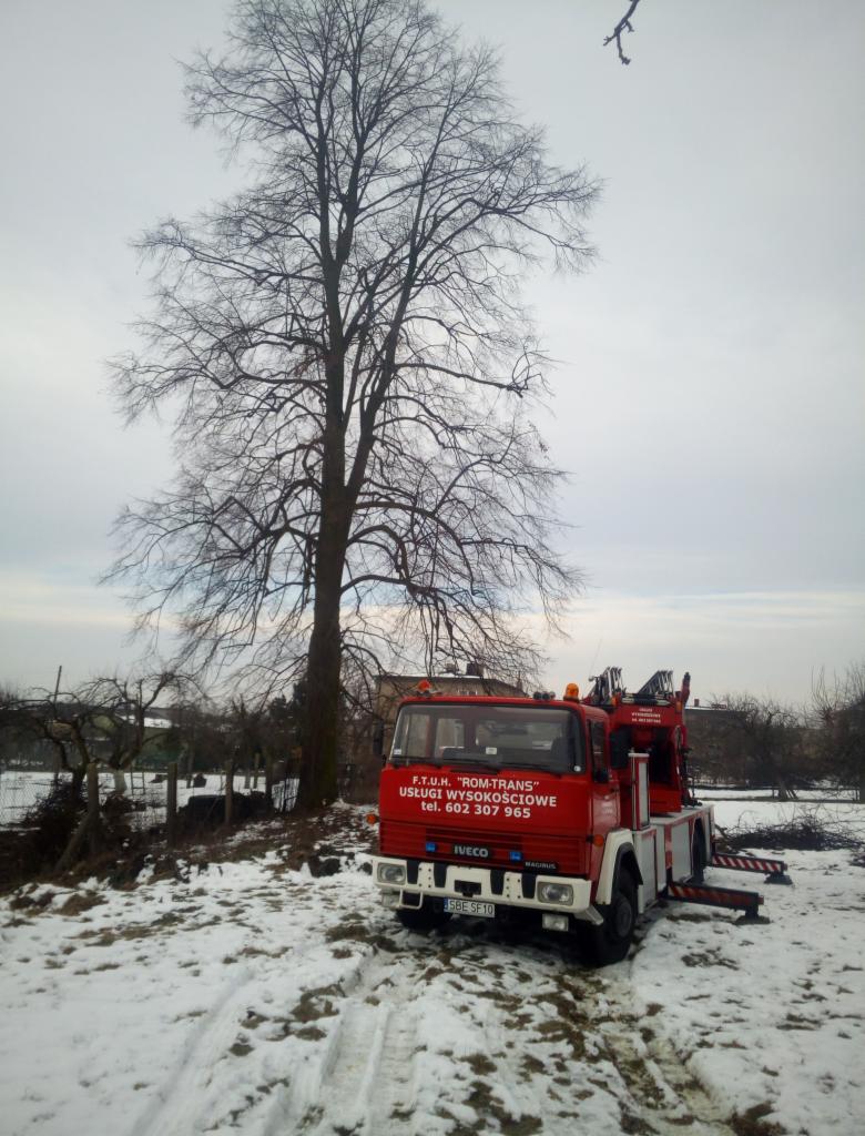 Czerwony samochód ciężarowy z podnośnikiem hydraulicznym firmy F.T.U.H. 'ROM-TRANS' zaparkowany na ośnieżonym terenie obok wysokiego, bezlistnego drzewa w pochmurny dzień.