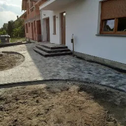 Fragment posesji z częściowo ułożoną szarą kostką brukową, widoczne przygotowane podłoże, schody wejściowe do budynku i fragment ściany budynku w tle.