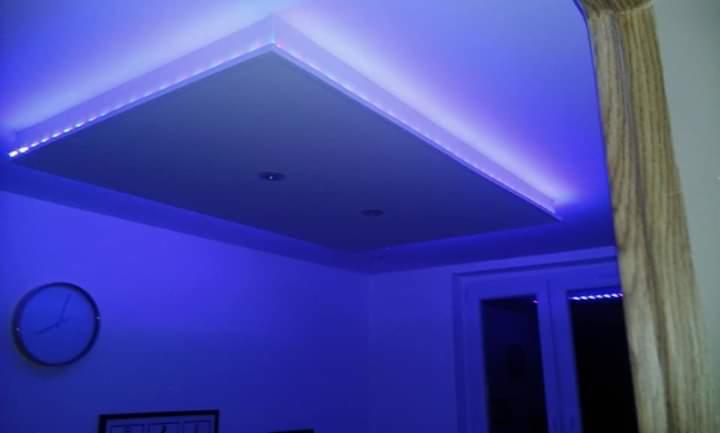 Podwieszany sufit z oświetleniem LED w kolorze niebieskim, widoczne dwa punkty świetlne, zegar na ścianie i fragment zasłony.