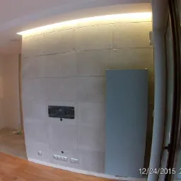 Wnętrze pokoju z drewnianą podłogą, szarą ścianą wyłożoną kwadratowymi płytami imitującymi beton, podświetleniem LED pod sufitem i dużym oknem balkonowym. Na ścianie widoczna puszka instalacyjna.