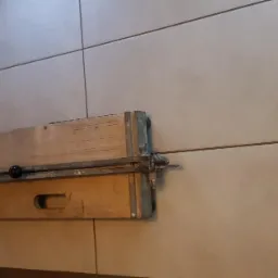 Ręczna przecinarka do płytek ceramicznych, przymocowana do ściany wyłożonej jasnymi, kwadratowymi płytkami. Widoczne ślady użytkowania na urządzeniu.