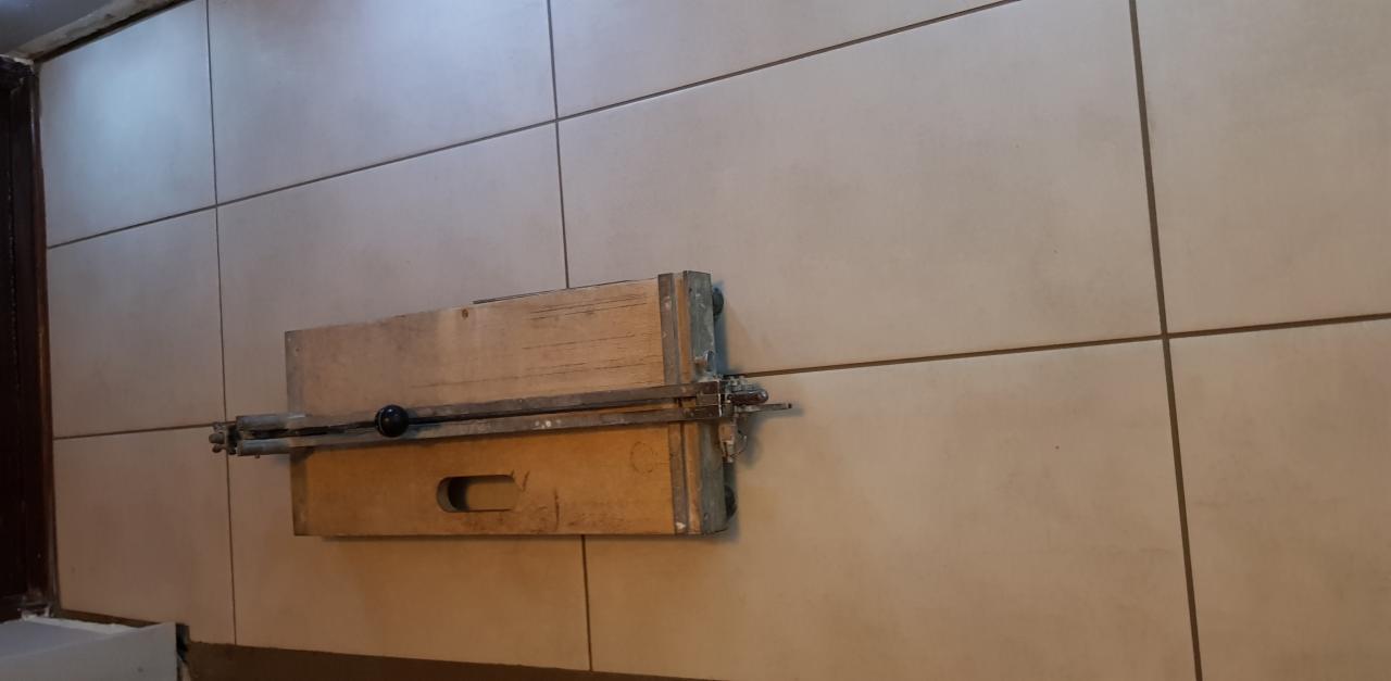 Ręczna przecinarka do płytek ceramicznych, przymocowana do ściany wyłożonej jasnymi, kwadratowymi płytkami. Widoczne ślady użytkowania na urządzeniu.