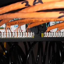 Panel krosowniczy z podłączonymi pomarańczowymi kablami sieciowymi CAT6, widoczne numery portów i oznaczenia kabli, czarne kable w tle.