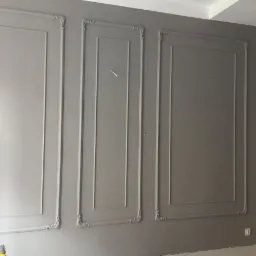 Szara ściana z trzema dekoracyjnymi panelami w stylu klasycznym, ozdobionymi sztukaterią w rogach, widok na fragment podłogi i drzwi.