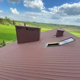 Brązowy, blaszany dach z kominem, wyłazem dachowym i wentylacją na tle zielonych pól i błękitnego nieba z chmurami.