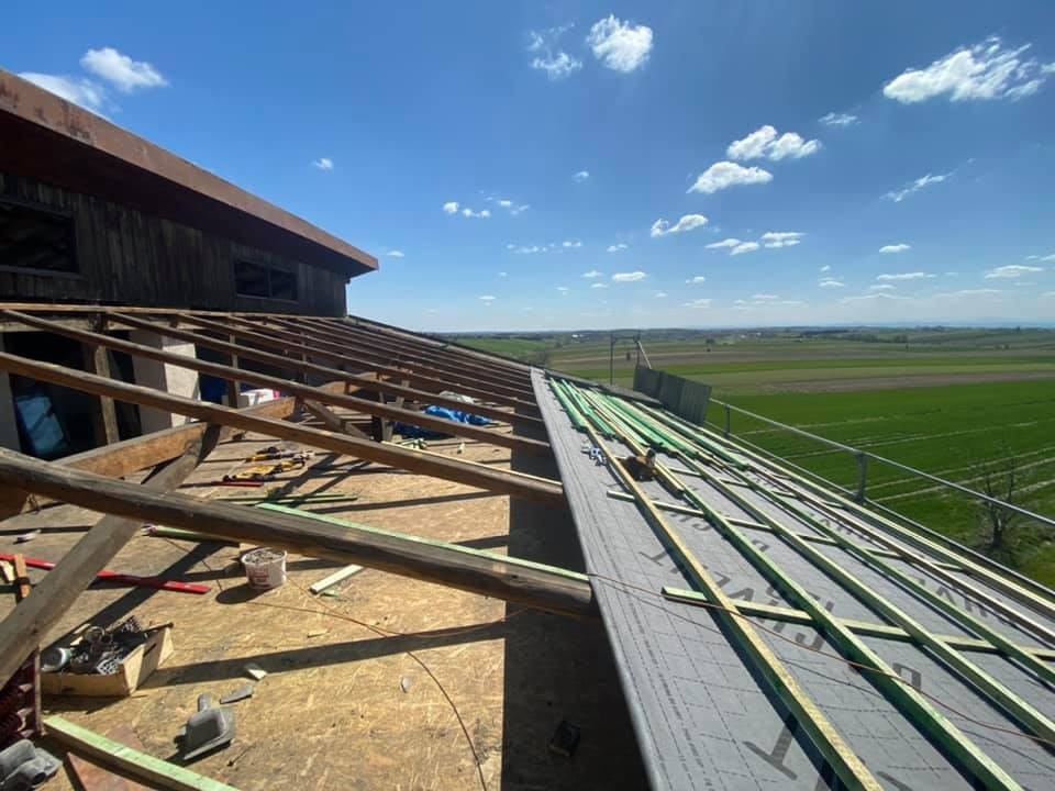 Konstrukcja drewniana dachu w trakcie montażu, widoczne elementy izolacji i łatowania na tle krajobrazu polnych łąk pod błękitnym niebem.