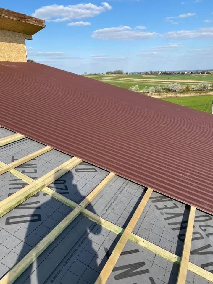 Częściowo ułożone brązowe blaszane pokrycie dachowe na łatach i membranie dachowej z widocznym krajobrazem w tle.