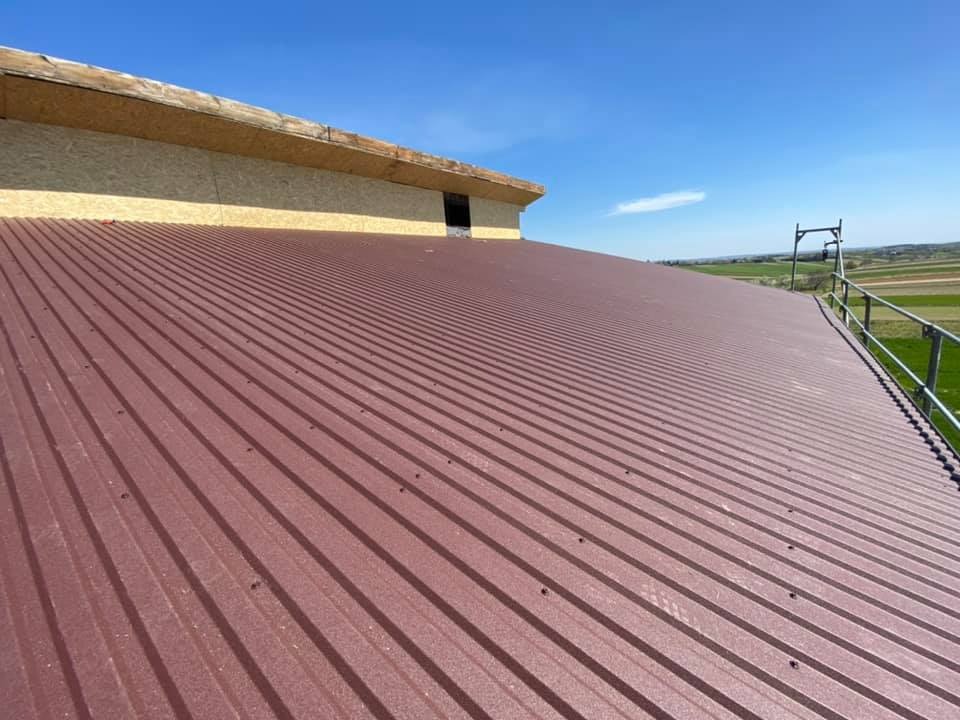 Nowy, brązowy dach z blachy trapezowej, widok z góry na fragment konstrukcji dachu z desek i niewielkie okno, w tle pola uprawne i błękitne niebo.