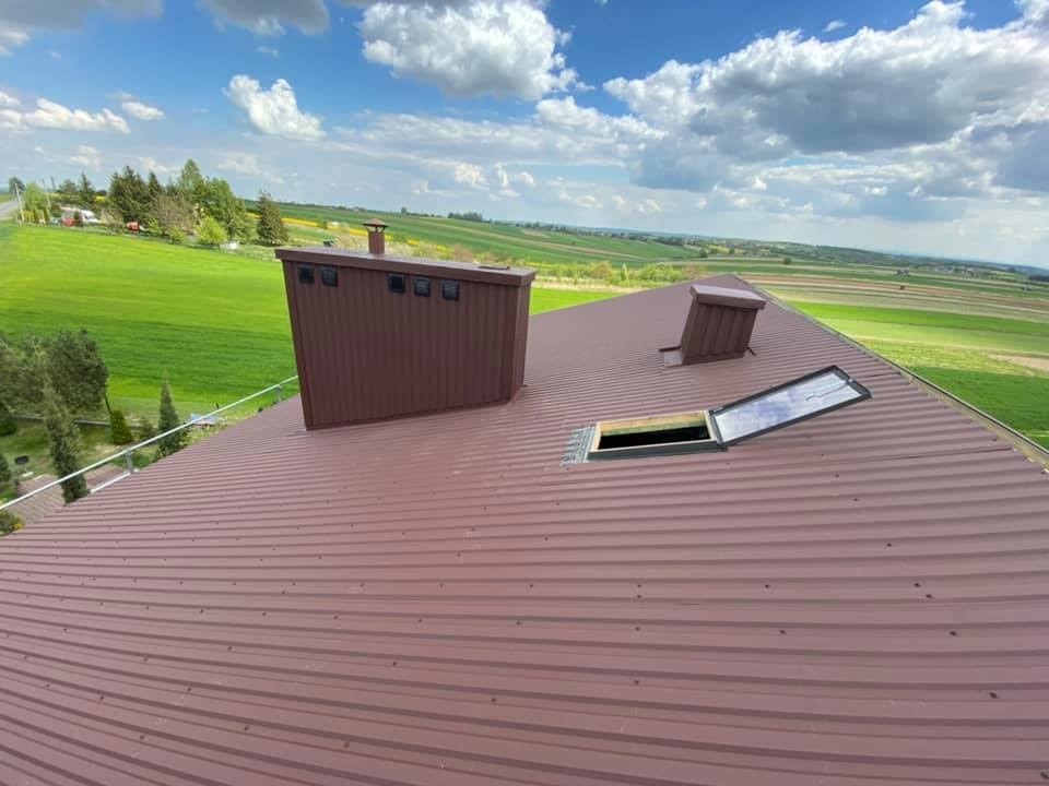 Brązowy, blaszany dach z kominem, wyłazem dachowym i wentylacją na tle zielonych pól i błękitnego nieba z chmurami.
