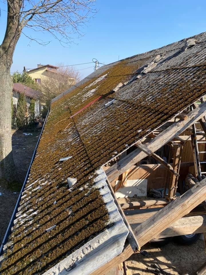 Stary dach z eternitu pokryty mchem, widoczne uszkodzenia i konstrukcja więźby dachowej, fragment rynny, w tle budynki mieszkalne i drzewo bez liści.