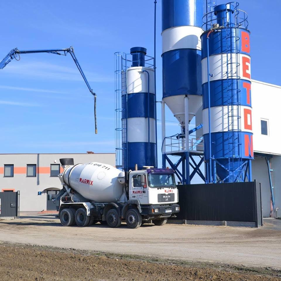 Betonomieszarka marki MAN na tle silosów betoniarni z napisem BETON i ramieniem pompy do betonu, w słoneczny dzień.