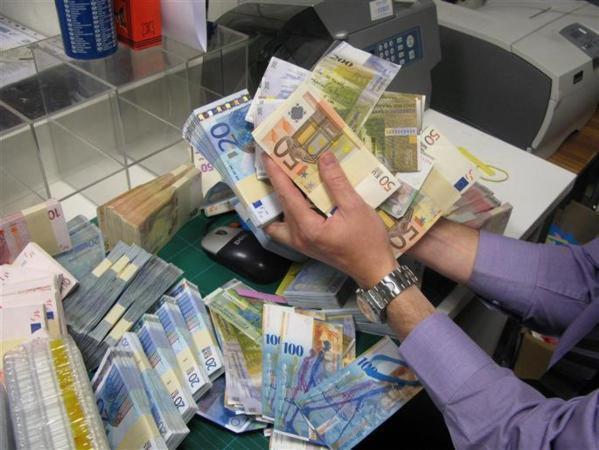 Stosy euro banknotów w różnych nominałach, trzymane w dłoniach osoby z zegarkiem na nadgarstku, na tle biurka z drukarką i innymi pakietami pieniędzy.