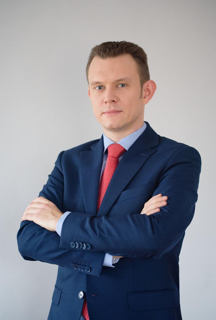 Mężczyzna w granatowym garniturze i czerwonym krawacie, z założonymi rękami, portret na jasnoszarym tle.