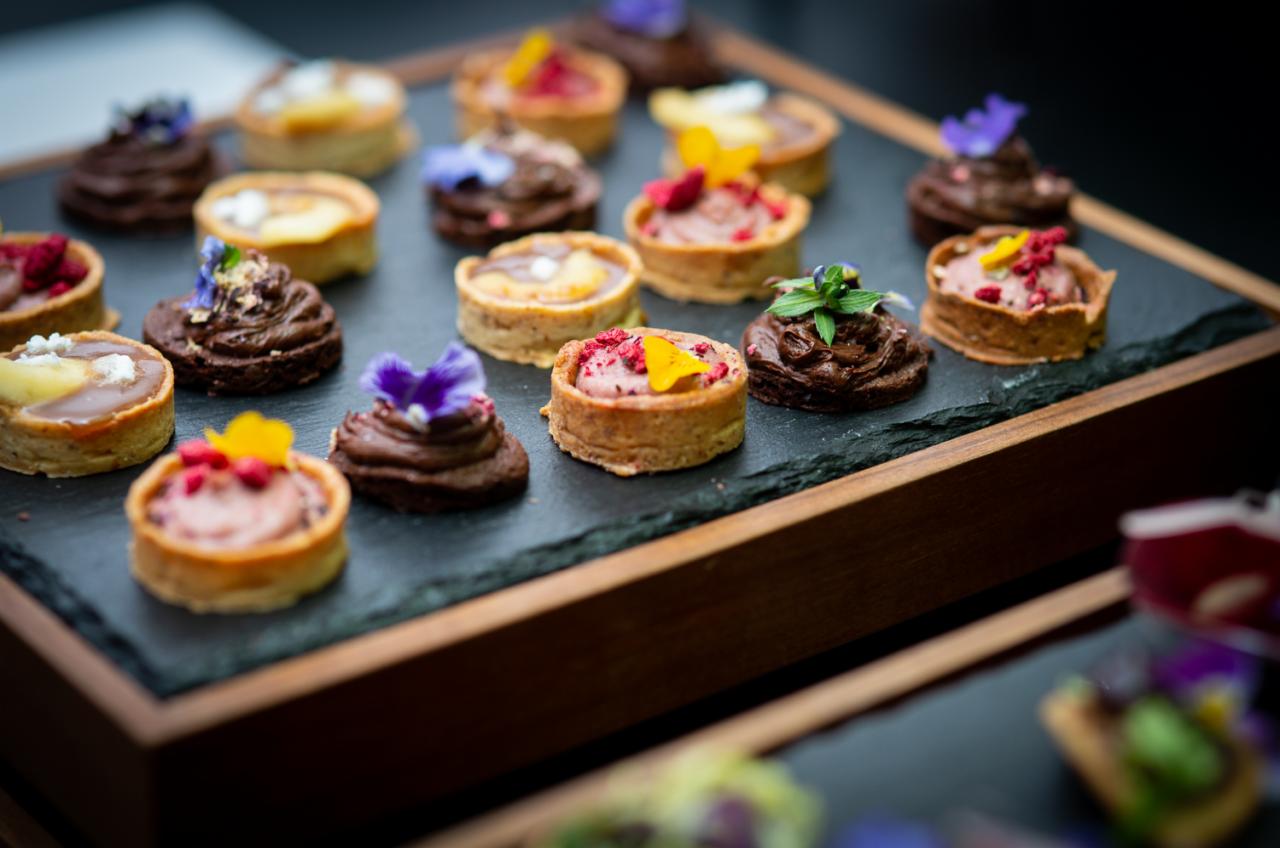 Miniaturowe tartaletki i czekoladowe babeczki udekorowane kwiatami i owocami, ułożone na czarnej, kamiennej tacy z drewnianą ramą.