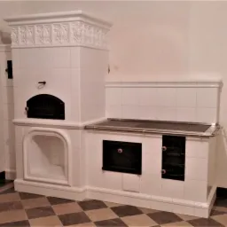 Biały, kaflowy piec kuchenny z czarnymi drzwiczkami piekarnika i płyty grzewczej, zdobiony reliefem, na szachownicowej podłodze.