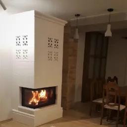 Biały, kaflowy piec kominkowy z paleniskiem, ozdobiony ażurowymi wzorami, płonący w salonie z drewnianą podłogą i koronkowym dywanikiem.
