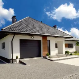 Dom jednorodzinny z ciemnoszarą dachówką karpiówką, białą elewacją i automatyczną bramą garażową w kolorze antracytu, na tle błękitnego nieba z chmurami.