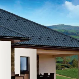 Czarny dach z dachówki ceramicznej na nowoczesnym domu z białymi ścianami i widokiem na zielone wzgórza w tle.