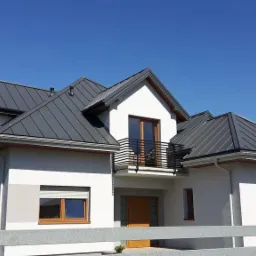 Nowoczesny dom jednorodzinny z ciemnoszarą blachą na dachu, widok z poziomu ogrodzenia, błękitne niebo w tle.
