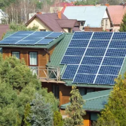 Dom jednorodzinny z drewnianą elewacją i panelami słonecznymi na zielonym dachu, widok zza drzew