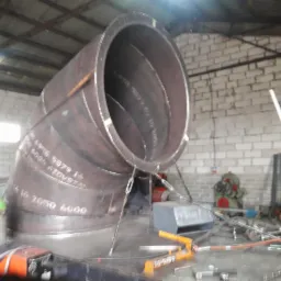 Duży, spawany kolanko rurociągu z metalu, podtrzymywany przez liny, widoczne napisy techniczne na powierzchni, w tle mur z białej cegły i narzędzia warsztatowe.