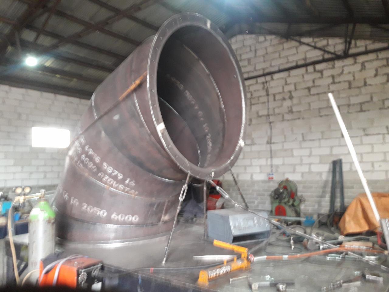 Duży, spawany kolanko rurociągu z metalu, podtrzymywany przez liny, widoczne napisy techniczne na powierzchni, w tle mur z białej cegły i narzędzia warsztatowe.