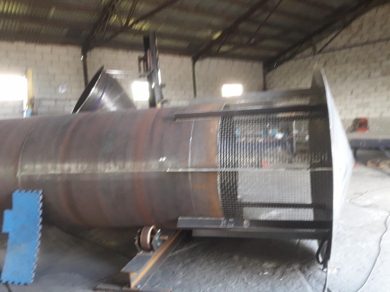 Częściowo zardzewiały, spawany zbiornik cylindryczny na rolkach, z kratownicą i stożkowym wlotem, w hali produkcyjnej.