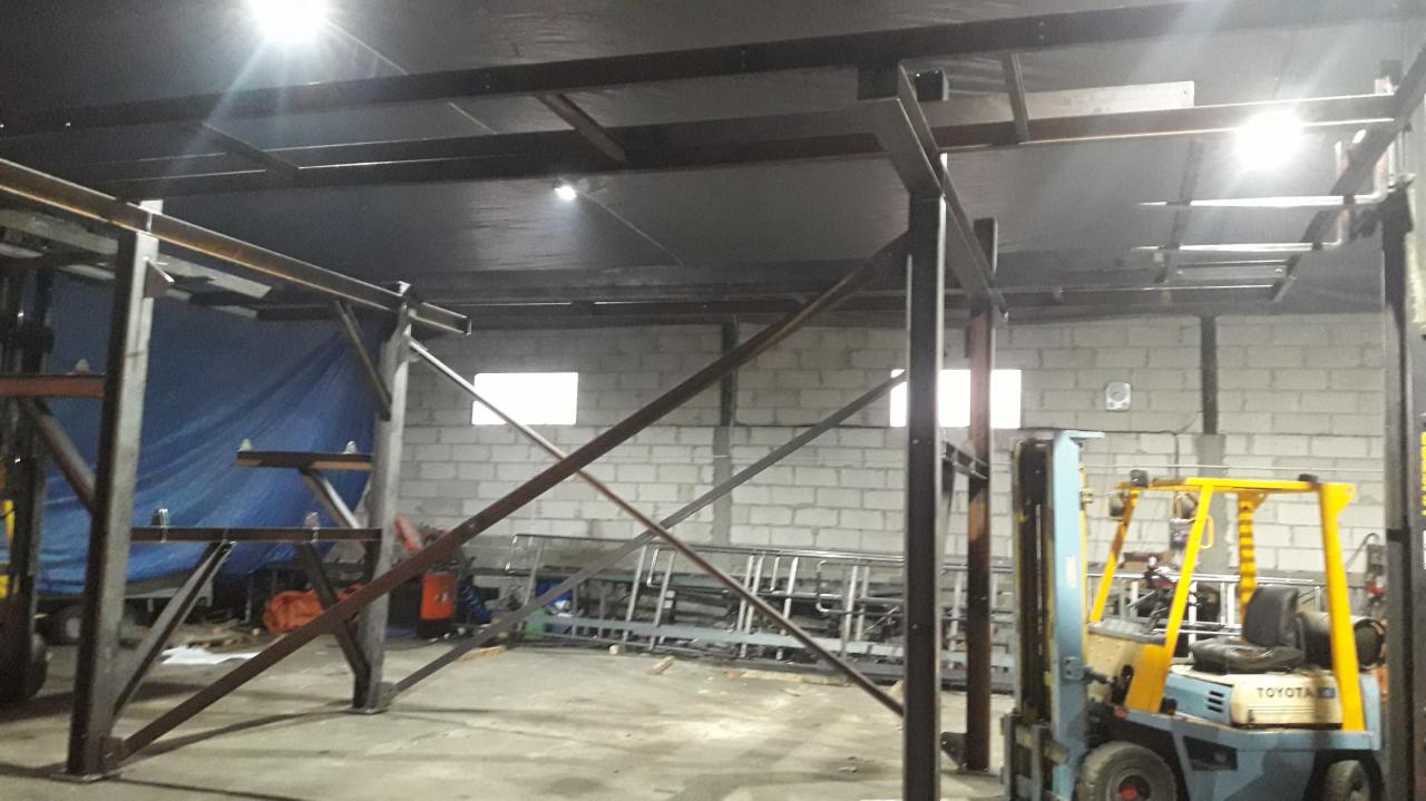 Konstrukcja stalowa podwieszana pod sufitem hali, widoczny wózek widłowy Toyota i ściana z białej cegły.