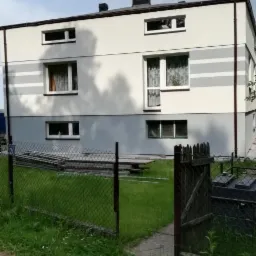 Dwukondygnacyjny budynek po odnowieniu elewacji, z szarymi poziomymi pasami, widoczny zza ogrodzenia z siatki i drewnianej furtki, na trawniku leżą elementy rusztowania.