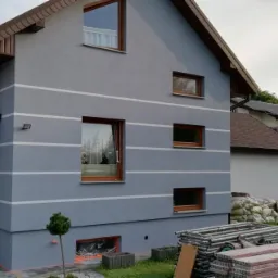 Szara elewacja domu jednorodzinnego z poziomymi białymi pasami, brązowe okna i dach, w tle widoczne rusztowania i worki z materiałem.