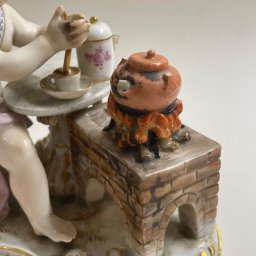 Natalia Kowalczuk Art - DA VINTAGE SPÓŁKA Z OGRANICZONĄ ODPOWIEDZIALNOŚCIĄ - Porcelanowa figurka przedstawiająca dziecko mieszające napój w filiżance przy stoliku, obok miniaturowy piec z garnkiem, całość zdobiona złotymi ornamentami.
