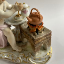 Natalia Kowalczuk Art - DA VINTAGE SPÓŁKA Z OGRANICZONĄ ODPOWIEDZIALNOŚCIĄ - Porcelanowa figurka przedstawiająca dziecko przy stoliku z filiżanką, obok czajnik na miniaturowej ceglanej kuchence z płomieniami, wszystko na zdobionej podstawie.