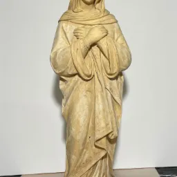 Stojąca, patynowana rzeźba Matki Boskiej z rękami złożonymi na piersi, na tle białej ściany.