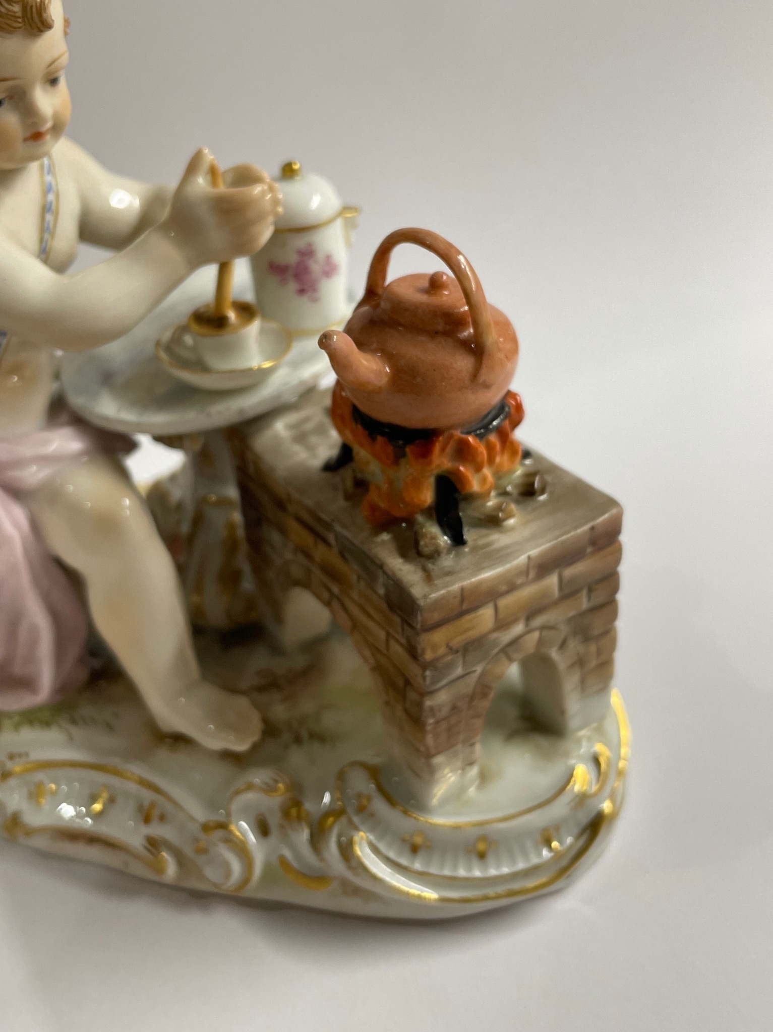 Porcelanowa figurka przedstawiająca dziecko przy stoliku z filiżanką, obok czajnik na miniaturowej ceglanej kuchence z płomieniami, wszystko na zdobionej podstawie.