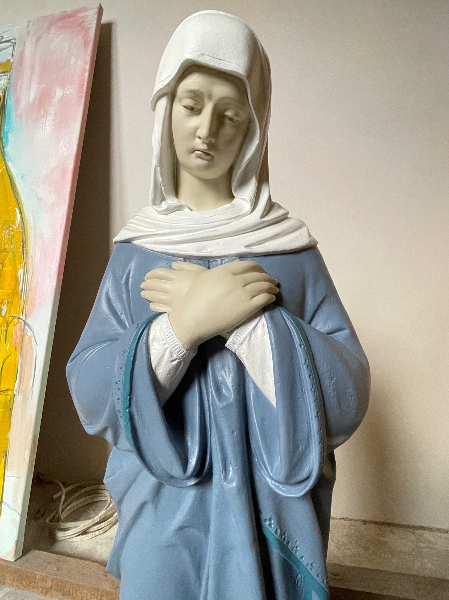 Statuetka Matki Boskiej w błękitnej szacie z białym nakryciem głowy, ręce skrzyżowane na piersi, obok niedokończony obraz na płótnie z elementami żółci i różu.