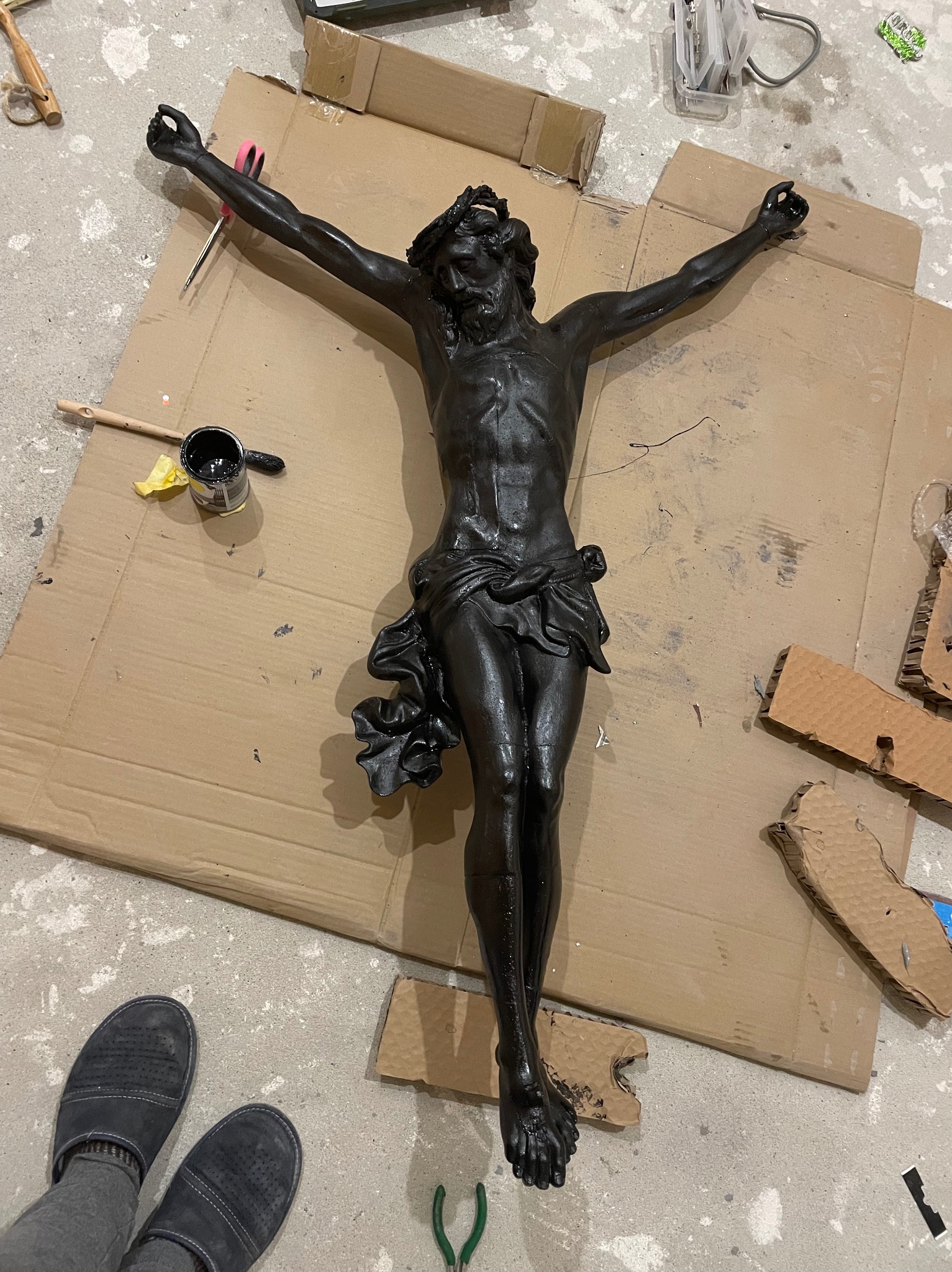 Czarna, pomalowana farbą figura Jezusa Chrystusa na kartonie podczas procesu renowacji, widoczne narzędzia malarskie, puszka z farbą i fragment obuwia osoby wykonującej pracę.