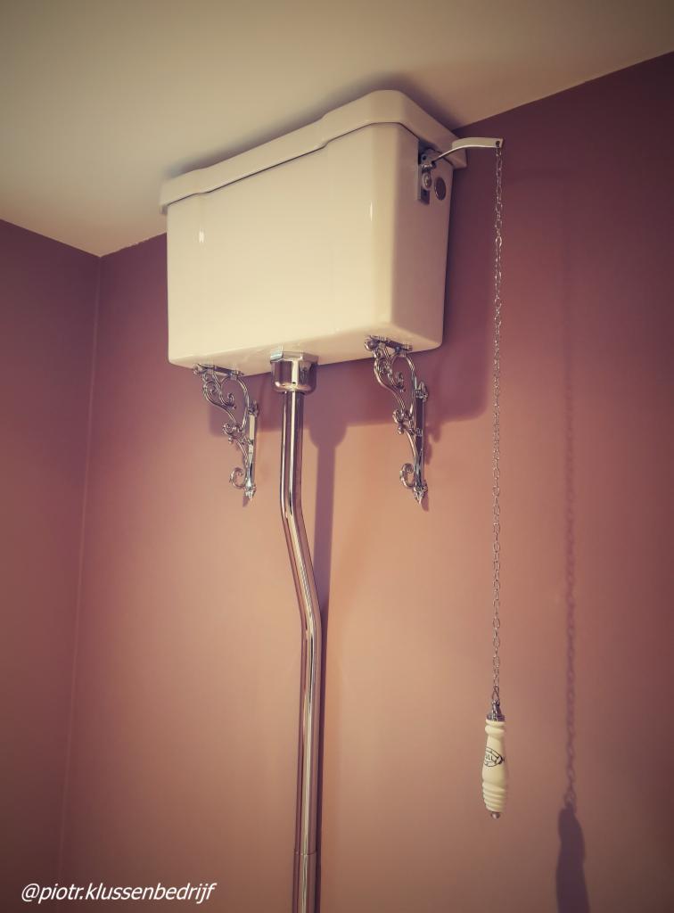 Zamontowana na ścianie spłuczka WC w stylu retro z ozdobnymi wspornikami i chromowaną rurą spustową na tle ściany w odcieniu pudrowego różu.