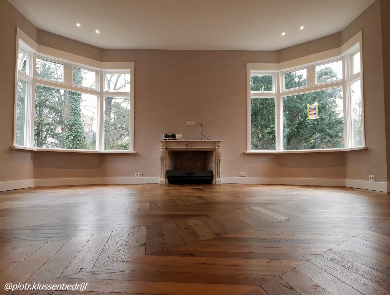 Jasne wnętrze po remoncie: parkiet ułożony we wzór jodełki francuskiej, ściany pokryte tapetą o delikatnej fakturze, dwa okna wykuszowe z widokiem na zieleń, dekoracyjny kominek.