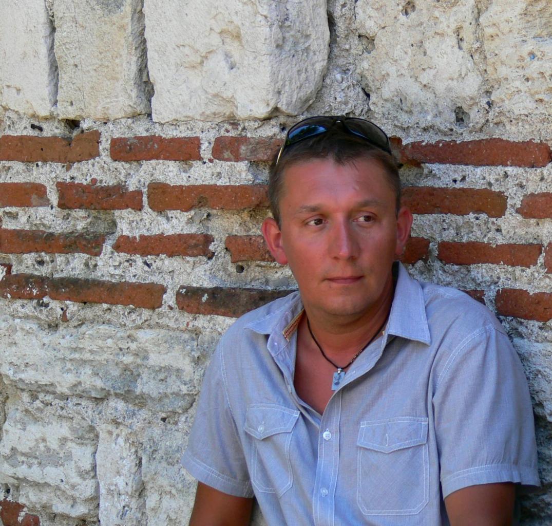 Mężczyzna w koszuli opiera się o starą ceglaną ścianę z kamiennymi elementami. Okulary przeciwsłoneczne na głowie, naszyjnik z zawieszką. Jasne światło dzienne.