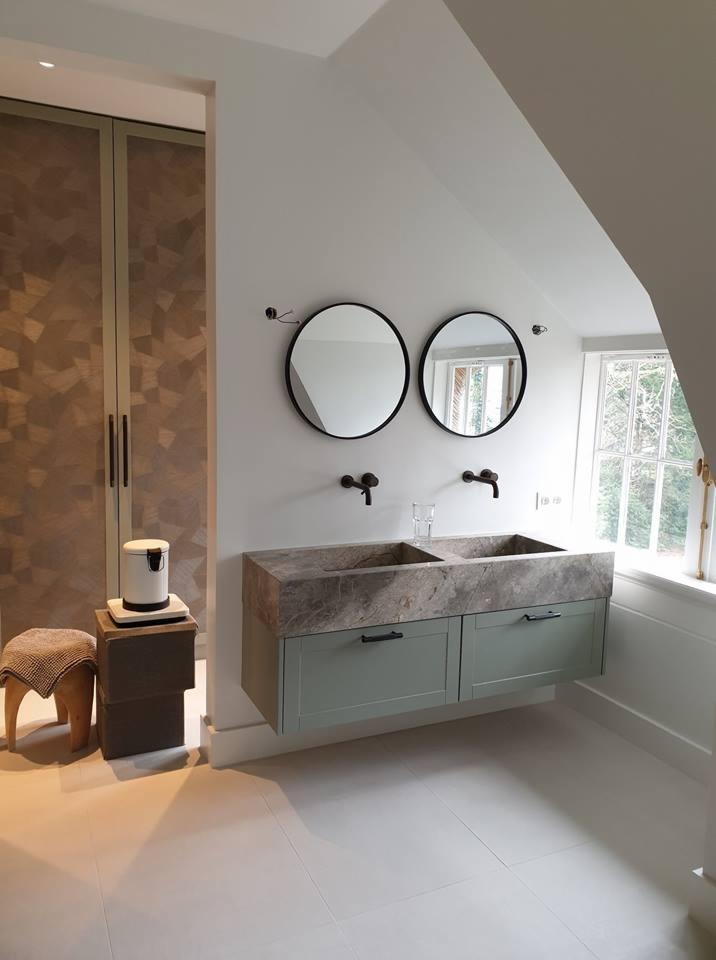 Nowoczesna łazienka w odcieniach szarości i zieleni, z podwójną umywalką z marmurowym blatem, okrągłymi lustrami i designerskimi bateriami, całość doświetlona naturalnym światłem z okna.