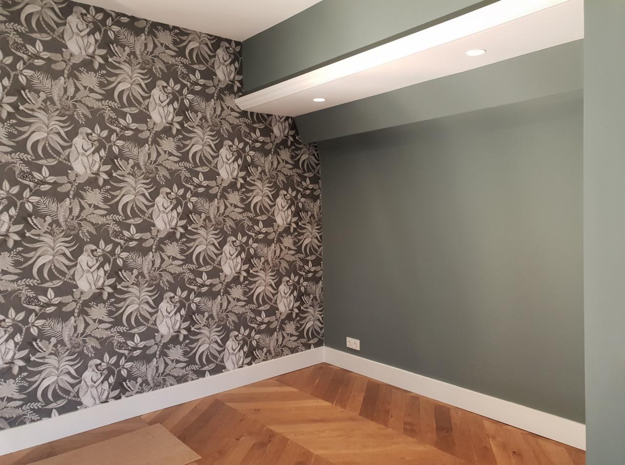 Pokój po remoncie: ściana pokryta tapetą w motywy roślinne i małpie, sąsiadująca z ścianą pomalowaną na oliwkowy kolor, parkiet ułożony w jodełkę, oświetlenie sufitowe.